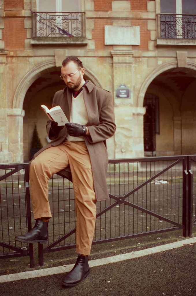Gabriel reading a book leaning on a railing at Place des Vosges, gloves and glasses.