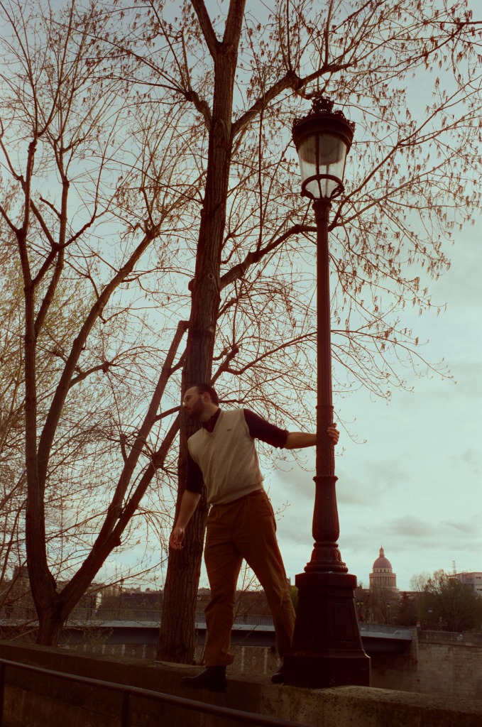 Gabriel perched on a low wall holding a street lamp, Île Saint-Louis quay at twilight.
