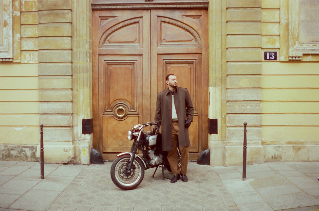 Gabriel, dandy in beige coat, leaning against a wooden porch in the Marais.