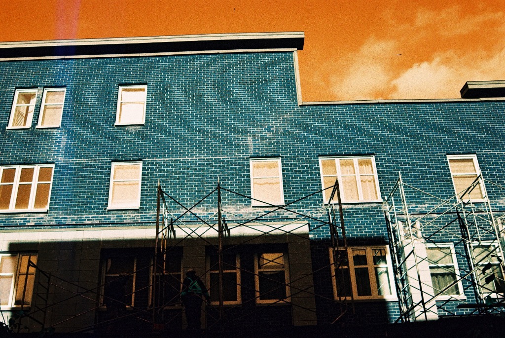 Red brick building turned blue-green with geometric architecture under orange sky.
