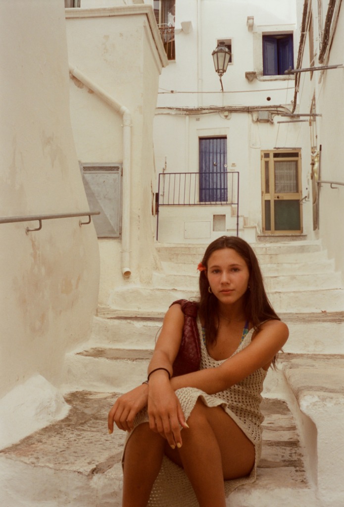 Young woman sitting cross-legged on white steps, brown dress.