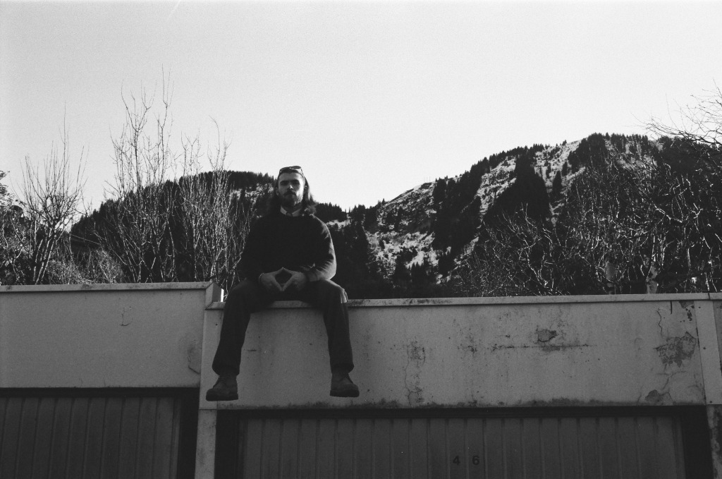 Man sitting legs dangling on a concrete roof, low angle view with mountains in background.