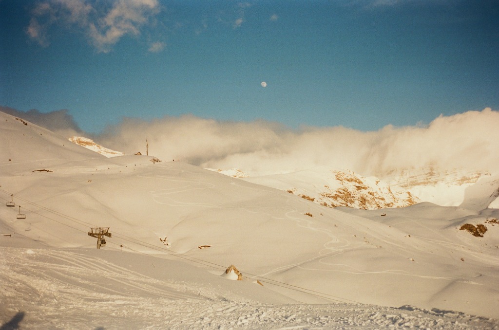 Moon visible in the sky above a snowy ridge golden by the setting sun.